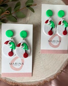 two earrings with green and red accents on top of a white card next to a plant