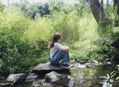 a young woman sitting on top of a rock next to a stream in the woods