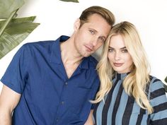 a man and woman standing next to each other in front of a white wall with plants