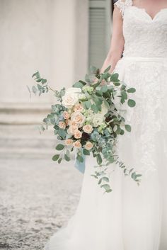 a bride holding a bouquet of flowers and greenery in front of an old building