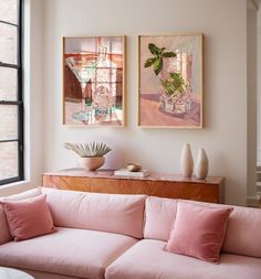 two paintings hang on the wall above a pink couch in front of a large window