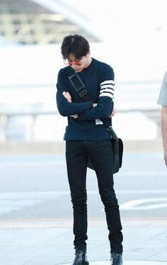 a man with his arms crossed standing in front of an airport terminal, wearing black pants and a blue sweater