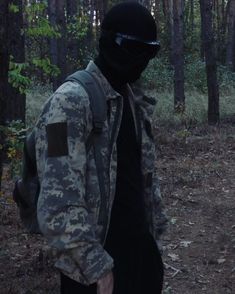 a man in camouflage jacket and goggles standing in the woods with his back to the camera