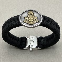 a black rope bracelet with a silver and gold masonic emblem on it, sitting on a white surface