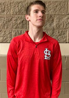 a young man standing in front of a brick wall wearing a red long sleeved shirt