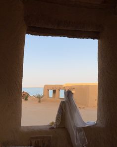 a person sitting on a ledge looking out at the water and sand, with a veil draped over their head