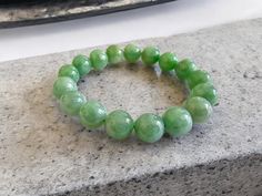 12mm each, 17 perfectly calibrated Green Burmese Jadeite Beads. This unique beads bracelet is 100% cut, calibrated, strung, and MADE IN JAPAN. Some of the rarest naturally occurring hues of A-Jadeite, calibrated Green creates a visually stunning Bracelet. With vibrant optics and translucency, the depth of each stone shines. This bracelet has light and darker green interwoven hues and tones, with marbling and fibrous optics. Completely untreated and 100% natural, this is a great accompaniment to formal and casual settings alike.  Each beaded-bracelet is one-of-a-kind, based on the natural variances of the raw jadeite, ensuring each one is truly unique and has its own story. Ref: A1IJGBJBB00005015 Materials: 51.50g Burmese Green A-Jadeite Dimensions: 12mm each Bead, 150mm stretch to 210 mm i Hand-strung Jade Stretch Bracelet With Round Beads, Jade Gemstone Beads Stretch Bracelet, Jade Stretch Bracelet With Gemstone Beads, Jade Stretch Bracelet With 8mm Beads, Green Stretch Bracelet With Polished Round Beads, Green Stretch Bracelet With Polished Beads, Green Board, Marbling, Jade Beads