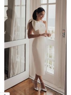 a woman standing in front of a window wearing a white dress and high heel shoes