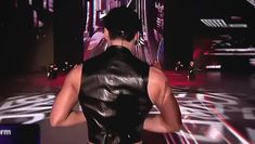 a man walking down a runway wearing a black leather vest and no shirt, with his back turned to the camera