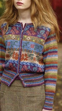 a woman with long red hair wearing a colorful cardigan sweater and skirt, standing in the woods