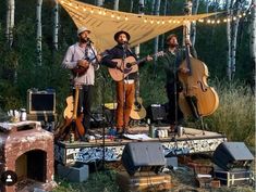 three men are playing guitars on a stage in the woods with string lights strung over them