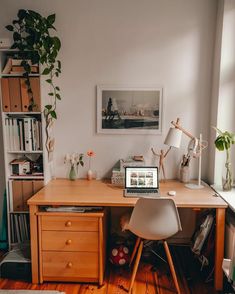 a wooden desk topped with a laptop computer next to a plant and bookshelf