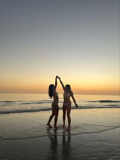 two women in bikinis standing on the beach at sunset with their hands up to each other