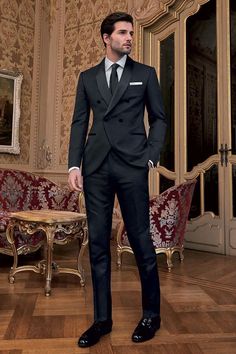 a man in a black suit and tie standing on a wooden floor next to a chair