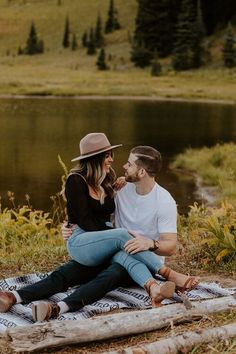 a man and woman sitting on a blanket in front of a lake while holding each other