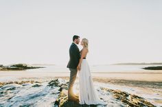 a bride and groom are standing on the rocks by the water at their beach wedding