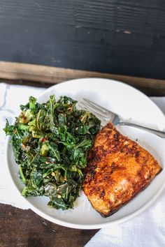 a white plate topped with meat and greens next to a fork on a wooden table