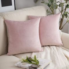 two pink pillows sitting on top of a white couch next to a book and potted plant