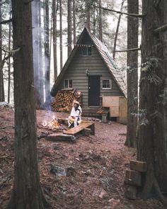 a woman sitting on a bench in the woods next to a fire pit and cabin