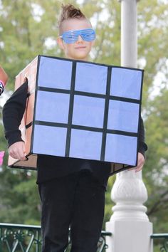 a young boy is dressed up as a rubik cube and wearing blue sunglasses while holding a cardboard box