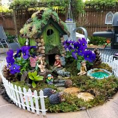 a fairy garden with purple flowers and gnomes in the yard, surrounded by a white picket fence