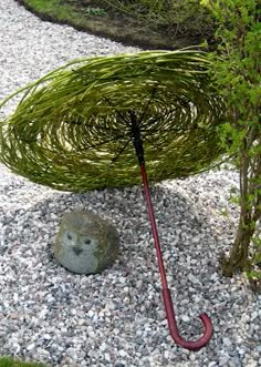 a red umbrella sitting on top of a gravel field next to a rock and tree