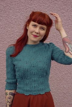 a woman with red hair wearing a green sweater and brown skirt, posing in front of a pink wall