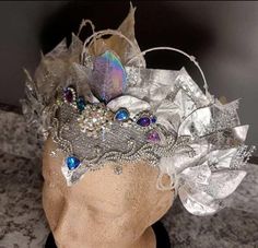 Enchanting Elegance: A Silver & Gray Headdress with Multicolored Gemstones Embrace the magic of color and style with this enchanting silver and gray headdress. Delicately adorned with shimmering foil leaves, this piece offers a touch of nature-inspired elegance. The headdress features a stunning array of blue, purple, and multicolored gems, each catching the light to create a dazzling display of brilliance. Intricately woven throughout are multicolored beads that add a playful pop of color and texture, bringing the entire design to life. Perfect for performances, festivals, or any occasion where you want to stand out, this headdress is a true statement piece, combining timeless sophistication with vibrant flair. Let your inner goddess shine with every step! Carnival Crown Headpiece For Mardi Gras, Crown Headpiece For Mardi Gras Carnival, Fantasy Headpieces For Mardi Gras Costume, Mardi Gras Carnival Crown Headpiece, Fantasy Costume Headpiece For Mardi Gras, Fantasy Mardi Gras Costume Hat, Fantasy Costume Hat For Mardi Gras, Mardi Gras Crown Headpiece For Costume, Mardi Gras Fantasy Crown Costume Headpiece