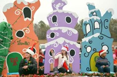 people sitting on the ground in front of some very colorful houses and trees with santa hats