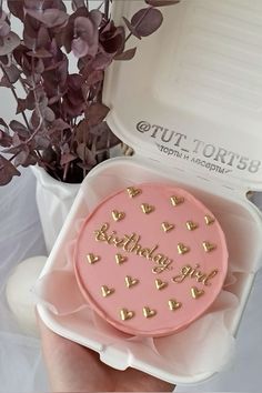 a pink birthday cake with gold hearts on it in a white container next to purple flowers