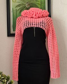 a woman wearing a pink crochet sweater and black dress standing next to a potted plant