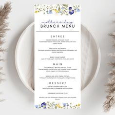 a white menu with blue and yellow flowers is on a plate next to some dried grass