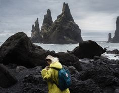 a person in a yellow jacket standing on rocks next to the ocean and looking at an island