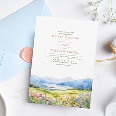 a wedding card with watercolor flowers and mountains in the background on top of a blue envelope