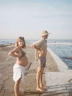 a pregnant woman standing next to a man on the beach with his hand on his hip