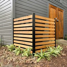 a wooden slatted fence in front of a building with plants and rocks around it