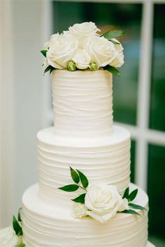 a wedding cake with white flowers on top and greenery around the edges, sitting in front of a window