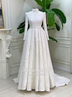 a white wedding gown with long sleeves and a high neckline is displayed in front of a plant