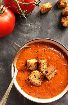 a bowl of tomato soup with croutons on the side and tomatoes in the background