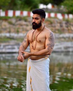 a man with a beard and tattoos standing in front of a body of water wearing a white robe