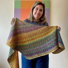 a woman holding up a multicolored crocheted shawl in front of a painting