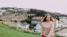 a woman with long red hair is walking on the grass near some houses and buildings