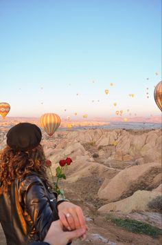 a woman is looking at hot air balloons flying in the sky above her and holding a rose