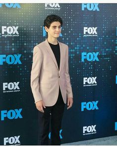 a young man in a pink suit and black shirt standing on a red carpet with his hands in his pockets