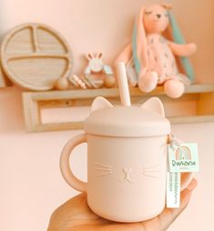 a hand holding a coffee cup with a cat on the inside and a stuffed animal behind it