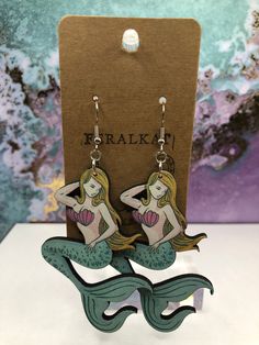 a pair of mermaid earrings sitting on top of a wooden stand next to a card