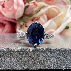 an oval blue sapphire and diamond ring sits on a table next to flowers in the background