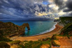 the beach is next to an arch shaped rock formation in the ocean under a cloudy sky