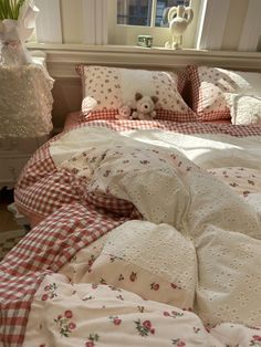 an unmade bed with red and white checkered bedspread, teddy bear on the pillow