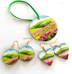 three pieces of art hanging from a green ribbon on a white surface, including earrings and pendants